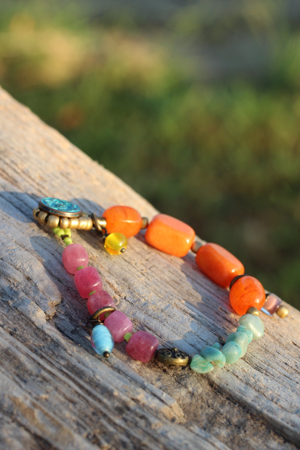A beaded bracelet with orange, pink, light blue, and mint green beads, displayed on a weathered wooden log with a blurred outdoor background.