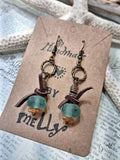 A pair of handmade dangle earrings displayed on a brown kraft card. Each earring has an antiqued bronze wire ring, a dark brown leather tie, a translucent light blue-green bead, and a small amber bead.