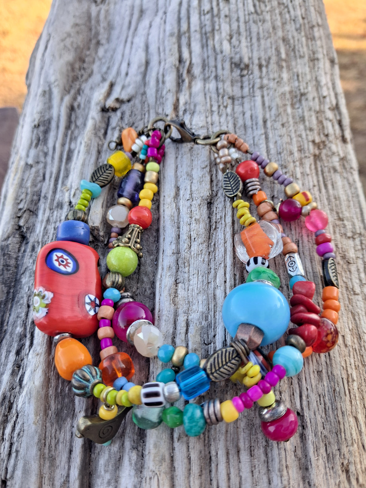 Handmade multi-strand bohemian beaded bracelet featuring colorful glass beads, a large red floral focal bead, round turquoise beads, seed beads, and brass charms resting on rustic weathered wood.