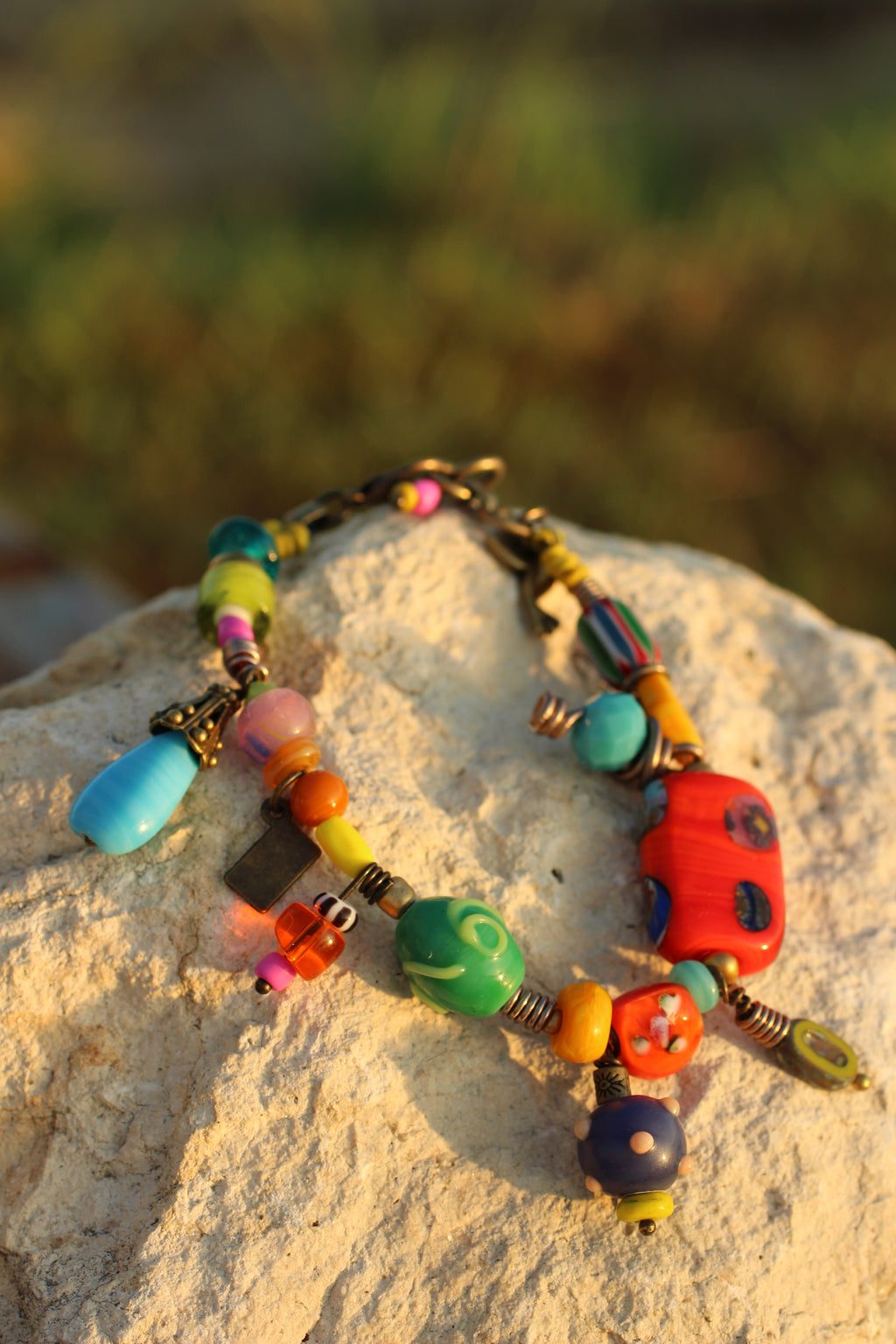 A colorful beaded necklace with various shaped glass-like beads in blue, green, red, orange, yellow, and pink, displayed on a light-colored stone with a blurred outdoor background.