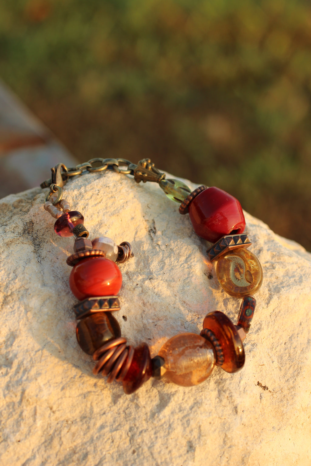 A beaded bracelet with red, amber, and brown glass beads and bronze-toned metal spacers, displayed on a light-colored stone with a blurred outdoor background.