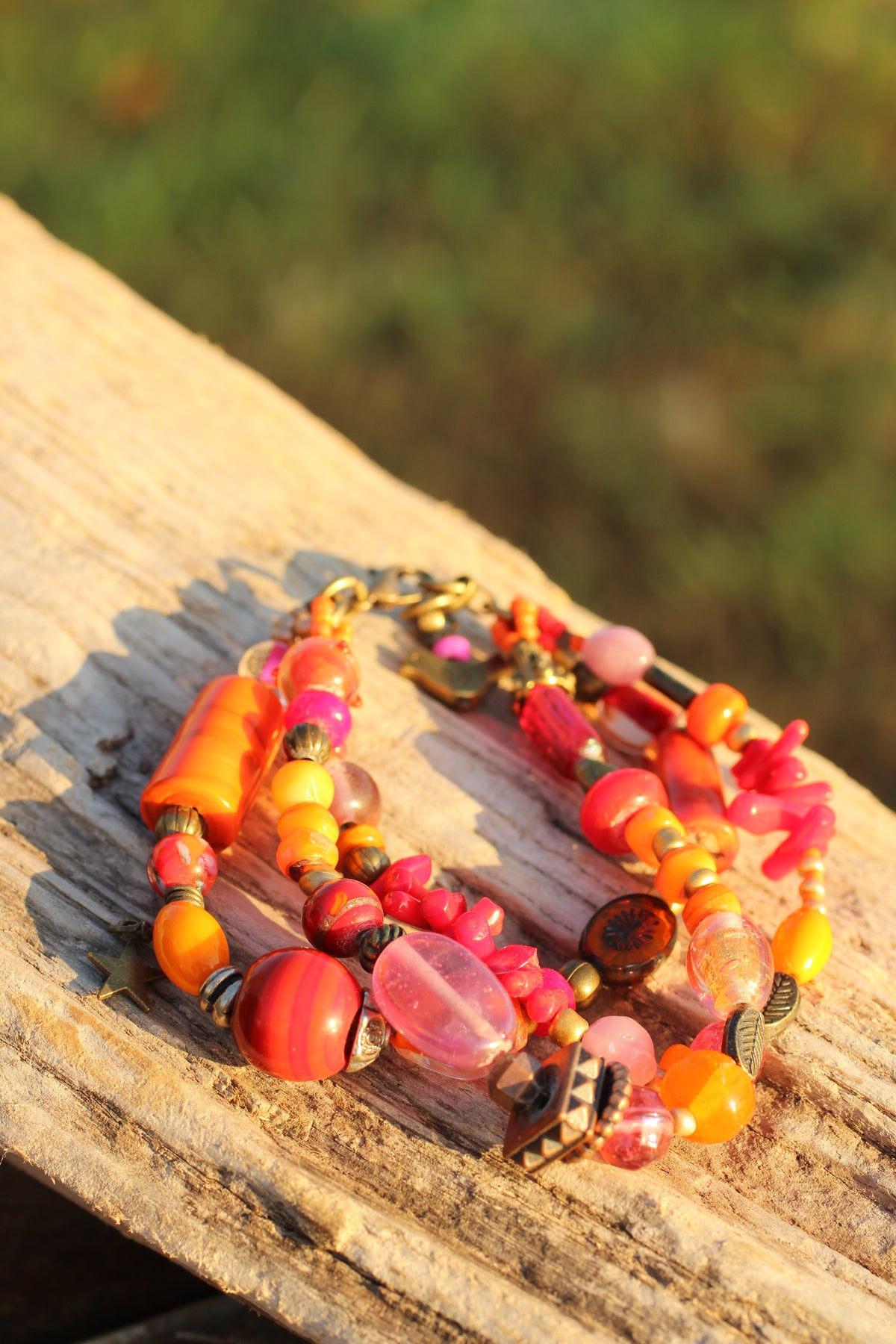 A close-up of a multi-strand beaded bracelet with vibrant orange, pink, red, and yellow beads, resting on a weathered wooden surface with a blurred green background.