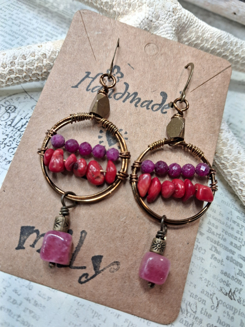 A pair of handmade bohemian hoop earrings with copper wire, pink and purple beads, and dangling pink stone charms, displayed on a kraft paper card.