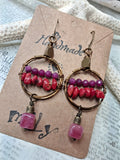 A pair of handmade bohemian hoop earrings with copper wire, pink and purple beads, and dangling pink stone charms, displayed on a kraft paper card.