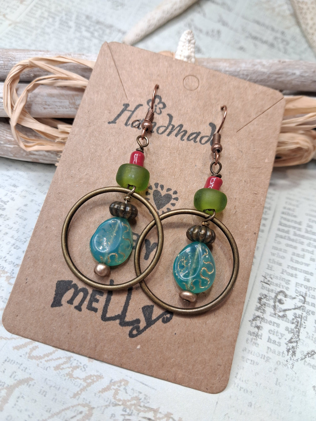 A pair of handmade dangle earrings displayed on a kraft paper card. The earrings feature bronze-toned metal hoops, green faceted beads, small red beads, and a prominent carved turquoise blue teardrop bead.