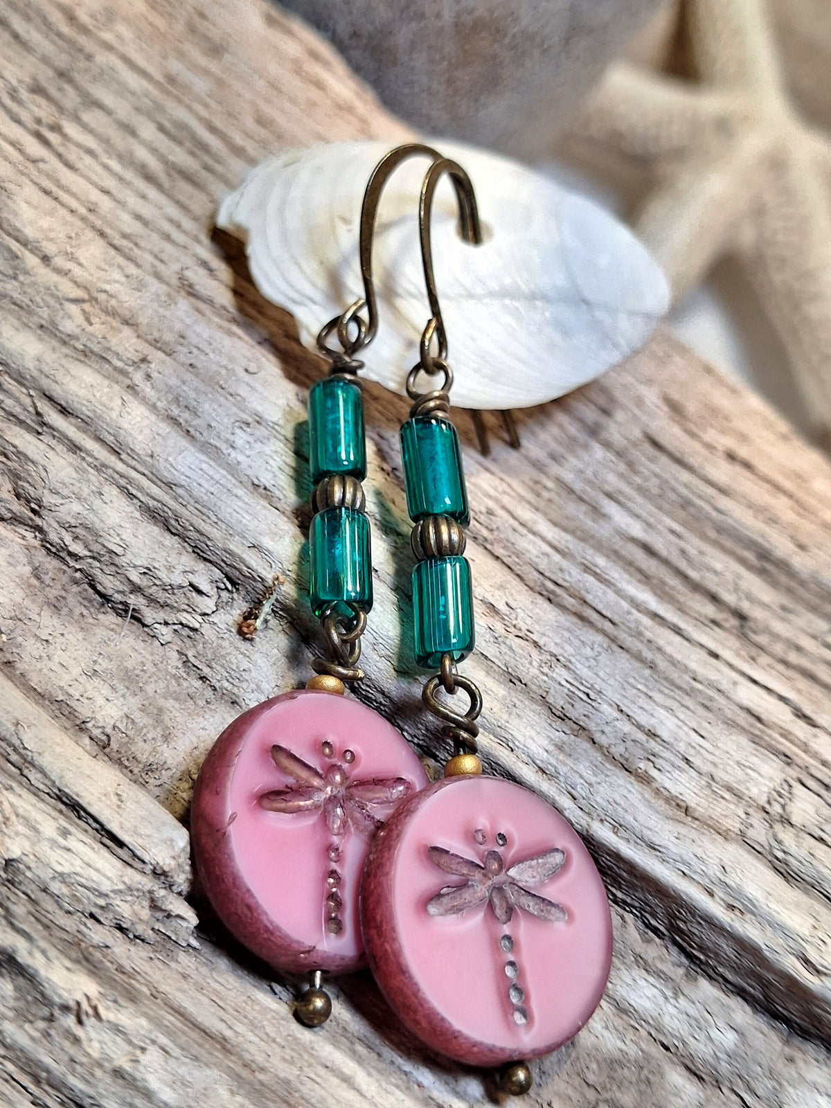 A pair of handmade dangle earrings with pink dragonfly charms, turquoise beads, and antique bronze hooks, displayed on a rustic wooden surface next to a white seashell.