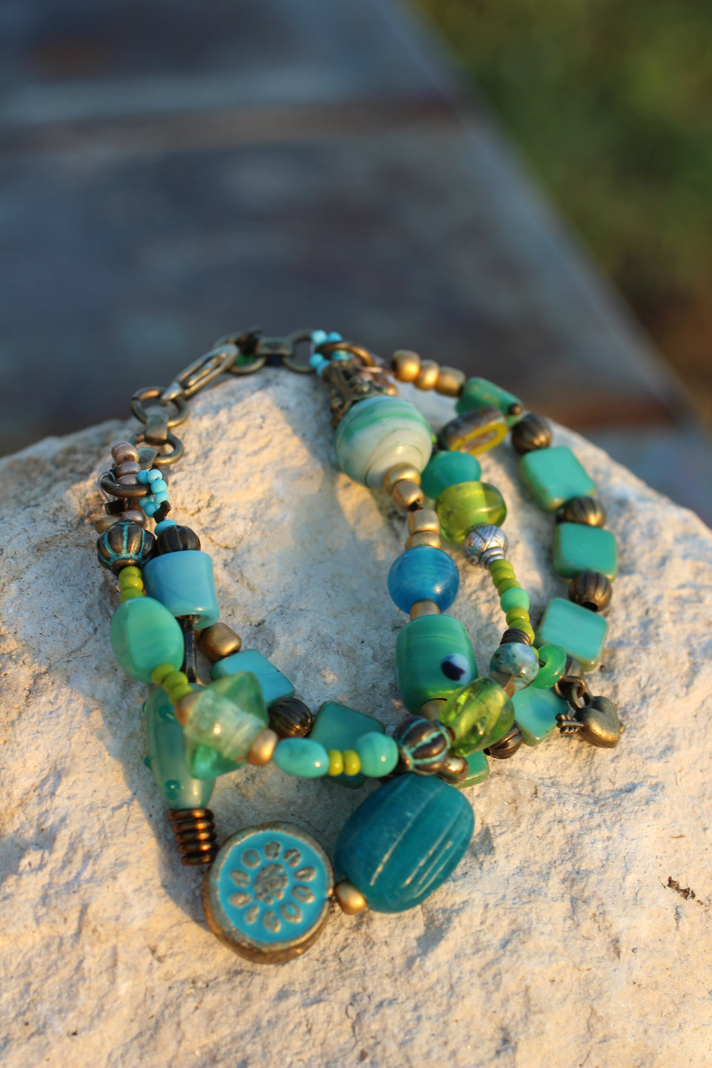 A multi-strand beaded necklace with turquoise, teal, and green beads, antique bronze-toned beads, and a round turquoise medallion pendant, displayed on a textured light stone surface outdoors.