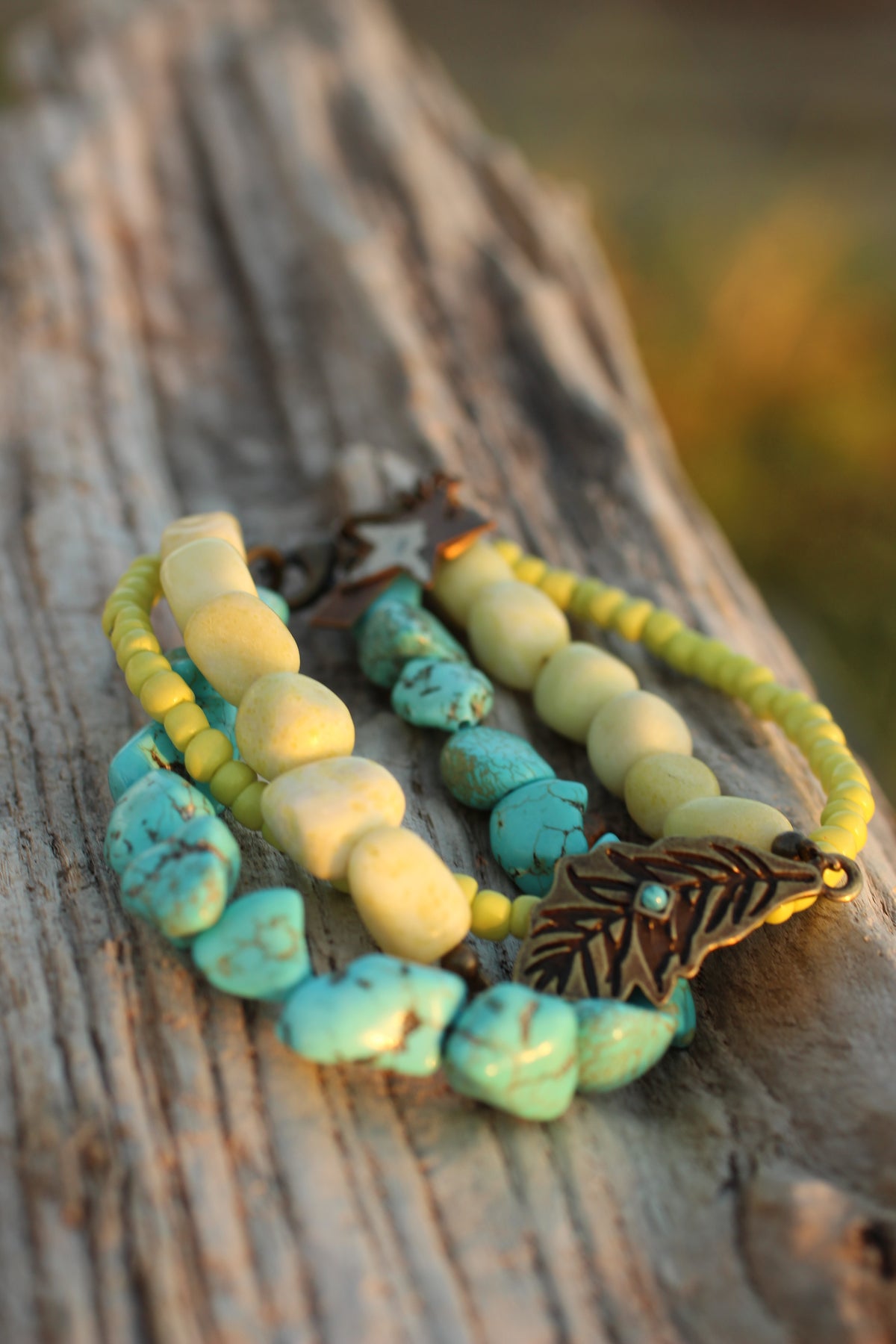 Close-up of a handmade multi-strand beaded bracelet featuring chunky turquoise stones, smooth pale yellow beads, small seed beads, and a bronze leaf charm, resting on weathered wood.
