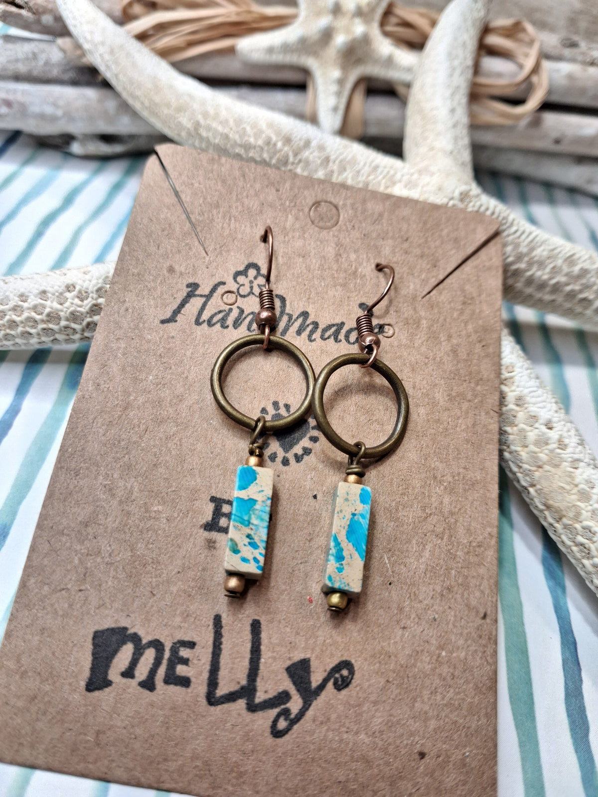 A pair of handmade dangle earrings displayed on a brown kraft card. The earrings feature bronze-toned metal rings, copper wire wrapped hooks, and rectangular turquoise and cream marbled impression jasper beads.