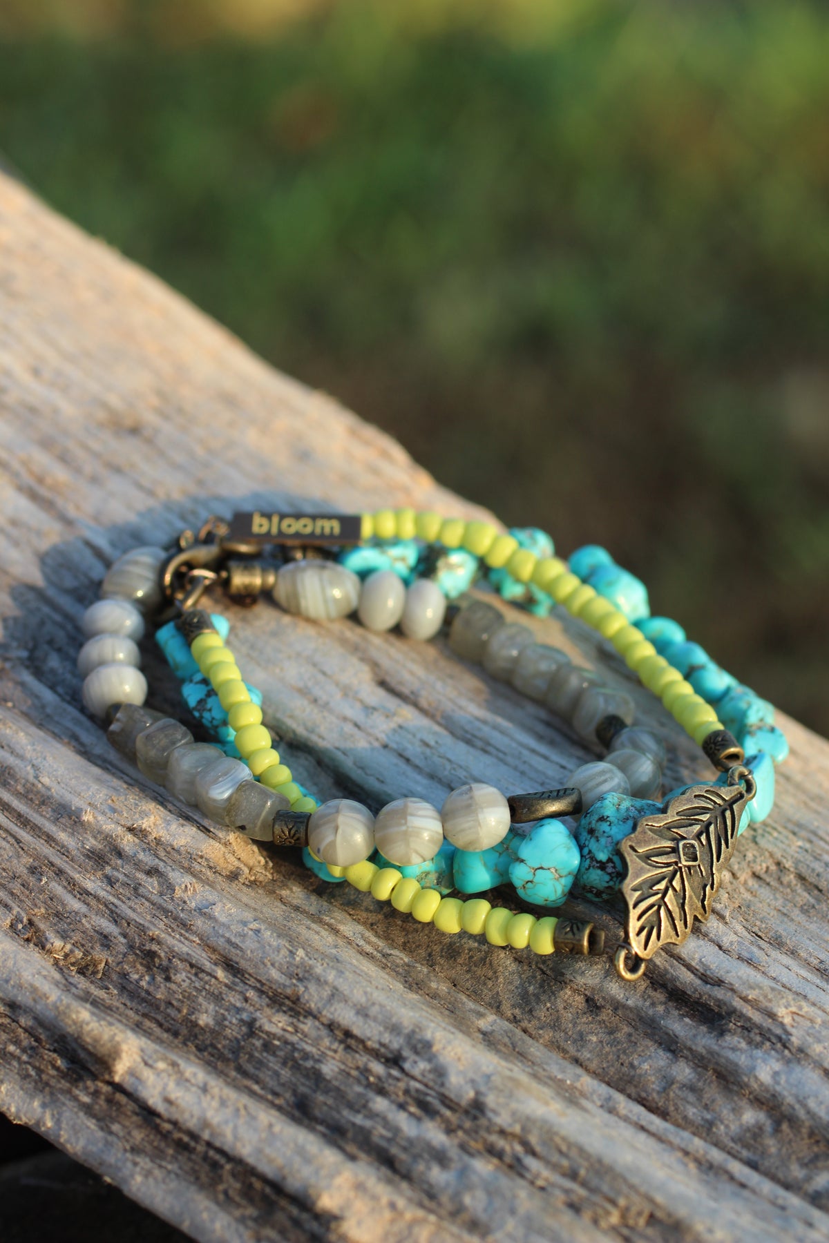 A set of three beaded bracelets with turquoise, lime green, and gray beads, featuring an antique bronze leaf charm and a clasp engraved with 'bloom', resting on a weathered wooden log with a blurred green background.