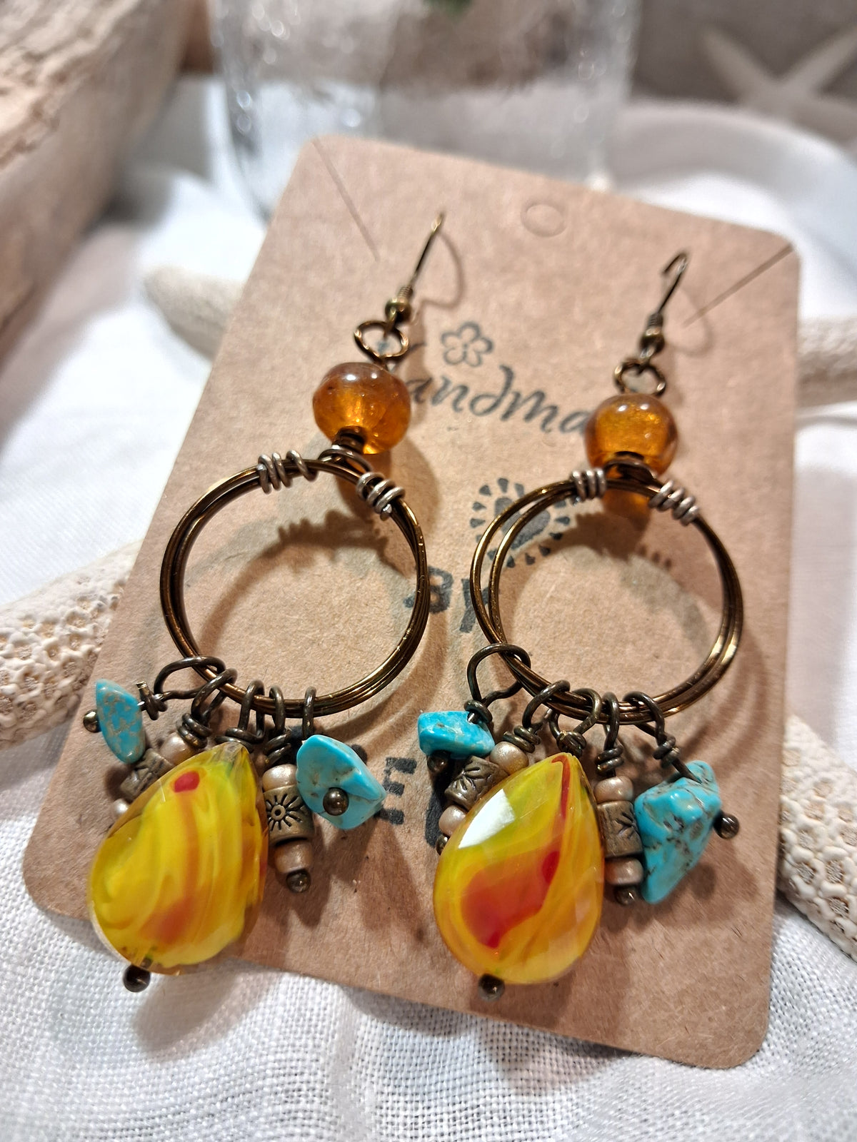 A pair of handmade dangle earrings displayed on a kraft paper card. The earrings feature antique bronze-toned metal hoops with twisted wire details, amber-colored beads, turquoise blue chips, and large marbled yellow and orange glass teardrop charms.