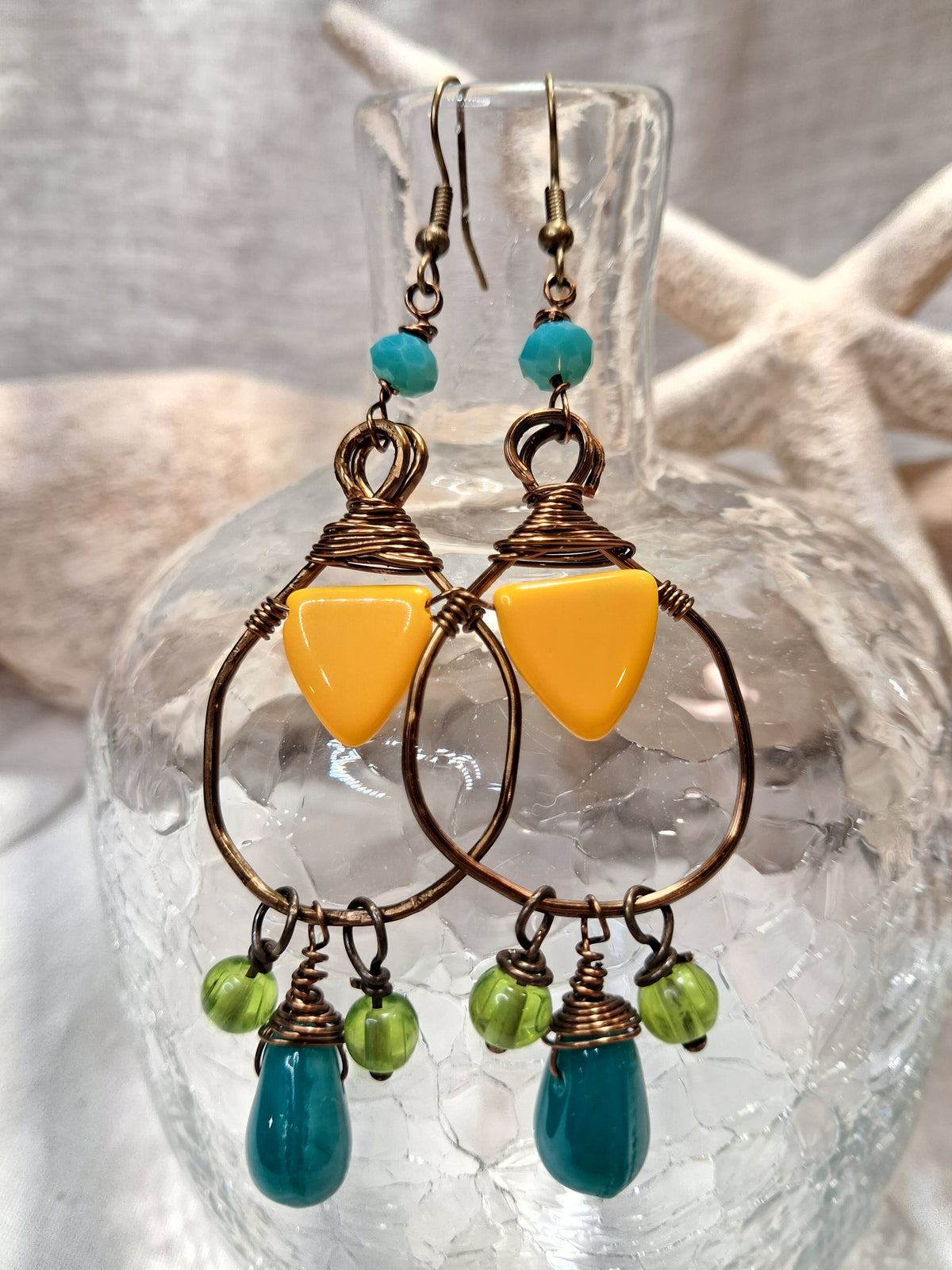 A pair of handmade dangle earrings displayed on a clear glass bottle. The earrings feature a large bronze hoop, a bright yellow triangular bead, small green beads, and a teardrop-shaped turquoise blue bead at the bottom.